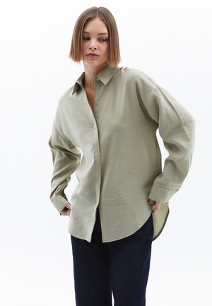 Chemise vert olive clair avec un devant à boutons, coupe oversize et manches longues. Présente un tissu texturé et un ourlet arrondi.
