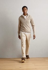 Light brown zip-up sweater over a white shirt, paired with light beige pants and brown shoes. The model stands on a wooden floor.
