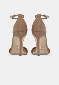 Nude korkokengät nilkkahihnoilla, sileää nahkaa, avonaista kärkeä ja hopeinen logo pohjassa. Stiletto-korko.
