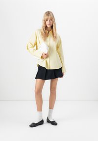 Gelbes Hemd mit Knöpfen, schwarzer plissierter Rock, weiße Clutch, graue Socken, schwarze Ballerinas. Einfaches Design mit lässiger Passform und weicher Textur.