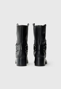 Botas negras de tobillo con cuero texturizado, tacones bajos y rectos, y correas decorativas con acentos metálicos. Vista trasera.