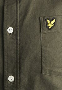 Camicia verde oliva con taschino, caratterizzata da una chiusura con bottone bianco e un logo di aquila nero e giallo ricamato sulla tasca. Tessuto standard.