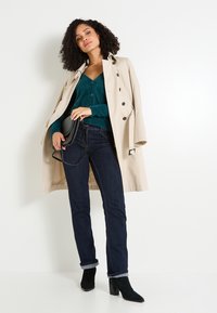 Trench beige, cárdigan de lana azul verdoso, jeans de mezclilla oscuros y botines negros. Modelo sosteniendo un pequeño bolso bandolera negro.