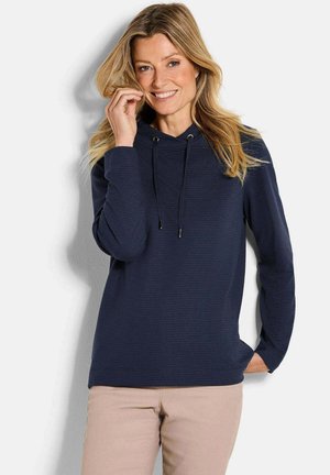 Marineblauwe hoodie met een textuurpatroon, trekkoordcapuchon en lange mouwen. Gecombineerd met lichtbeige broek tegen een neutrale achtergrond.