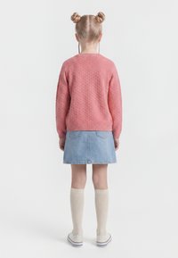 Fiatal lány szemből háttal áll, rózsaszín pulóvert, világoskék farmer szoknyát, térdig érő krémszínű zoknikat, fehér tornacipőt visel, és két kontyban hordja a haját.