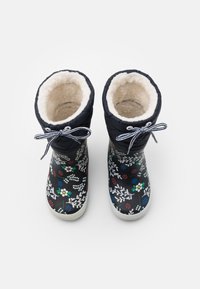 Bottes étanches de couleur marine avec doublure en fausse fourrure blanche, arborant un motif floral rouge, bleu et blanc. Agrémentées d'un ruban rayé sur le dessus.