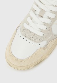 Αθλητικό παπούτσι από μπεζ σουέτ και λευκό δέρμα, με υφή στη μύτη, λευκά επίπεδα κορδόνια, μικρές οπές στη μύτη και μπεζ λαστιχένια σόλα.