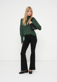 Un pull à col roulé vert côtelé, un jean noir évasé et des ballerines noires. Le mannequin tient un petit sac à main noir. Fond blanc uni.