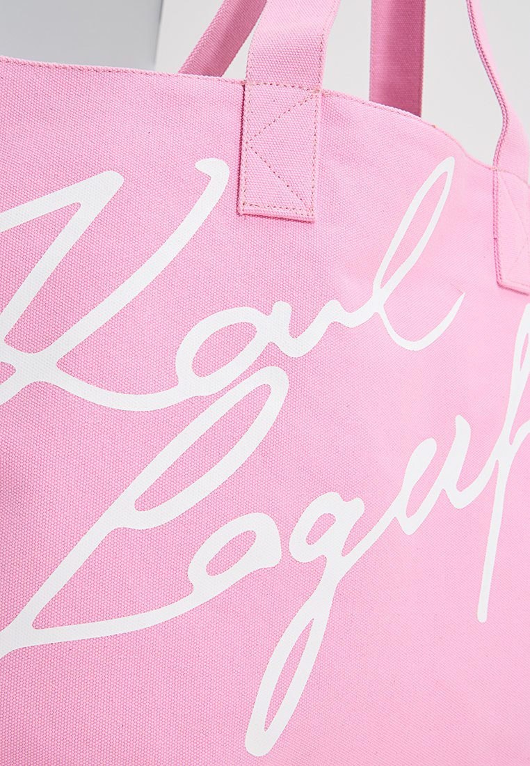 Sac en toile rose avec le logo "Karl Lagerfeld" en cursive blanche et des anses renforcées sur fond gris clair.