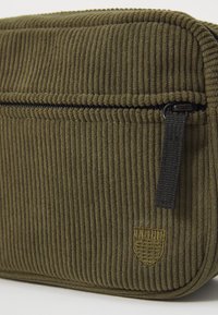 Oliven grøn corduroy taske med en struktureret overflade, lynlåslukning og et lille broderet logo i nederste hjørne. Sorte lynlås detaljer.