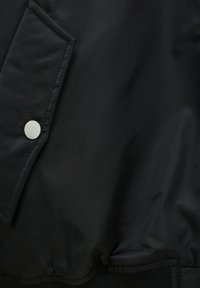 Veste en tissu noir en gros plan montrant une poche à rabat avec un bouton-pression argenté et un détail élastiqué à l'ourlet.