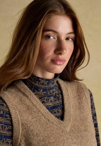 Pruun kudumiveste V-dekoltee ja tumesinise kõrge kaelusega särk, millel on kuldne ratsaspordi muster. Sile tekstuur ja kihiline disain.