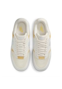Zapatillas Nike con una parte superior de malla blanca, acentos de ante amarillo y una mediasuela texturizada. Presenta cordones color crema y un logo gris en los laterales.