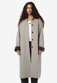 Vrouw met lang krullend haar, gekleed in een beige lange jas met zwarte kraag en manchetten over een witte top en zwarte broek.