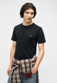 Νεαρός άνδρας με σκούρα μαλλιά φοράει μαύρο t-shirt με ένα μικρό λογότυπο άλκη και ένα καρό πουλόβερ δεμένο γύρω από τη μέση του, στέκεται με το ένα χέρι στην τσέπη.