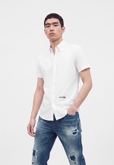 Jeune homme portant une chemise blanche à manches courtes et un jean bleu usé, debout avec une main dans la poche.