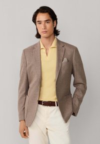 Man draagt een beige blazer met een patroon zakdoek, een gele overhemd met kraag, een bruine geweven riem en crèmekleurige broek, staand tegen een grijze achtergrond.