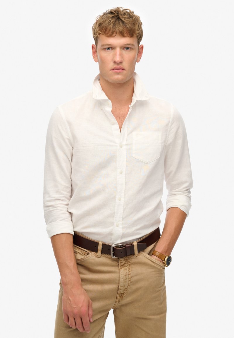 Camisa blanca de lino con botones, mangas remangadas y un bolsillo en el pecho, combinada con pantalones beige y un cinturón y reloj de cuero marrón.