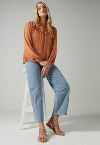 Rust-colored blouse with ruffle detail, button front, and long sleeves; paired with light blue straight-leg jeans and tan sandals.