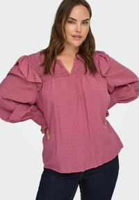 Kobieta z długimi brązowymi włosami, ubrana w różową bluzkę o teksturowanym wzorze z bufiastymi długimi rękawami i ciemnoniebieskimi dżinsami, z rękami na biodrach, na jasnym tle.