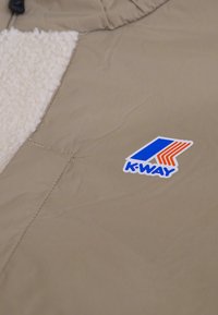 Giacca beige con un pannello in pile bianco testurizzato. Presenta un logo "K-WAY" colorato e una chiusura con zip dotata di cursore sulla colletto.