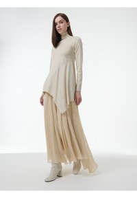 Beige langemouwen top met een hoge hals en asymmetrische zoom, gecombineerd met een vloeiende, vloerlange beige rok. Witte hakken ronden de look af.