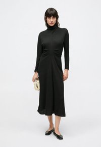 Μαύρο μίντι φόρεμα με μακριά μανίκια και ραβδώσεις, με ψηλό λαιμό και μια στριφτή λεπτομέρεια στη μέση. Συνδυάζεται με μαύρα φλατ παπούτσια και κρεμ τσάντα χειρός.