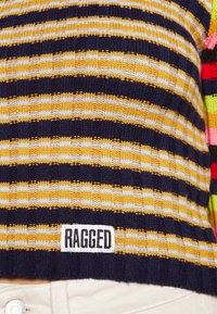 Maglione lavorato a maglia con strisce orizzontali gialle, blu navy e bianche, caratterizzato da un'etichetta "RAGGED" vicino all'orlo inferiore sopra pantaloni bianchi.