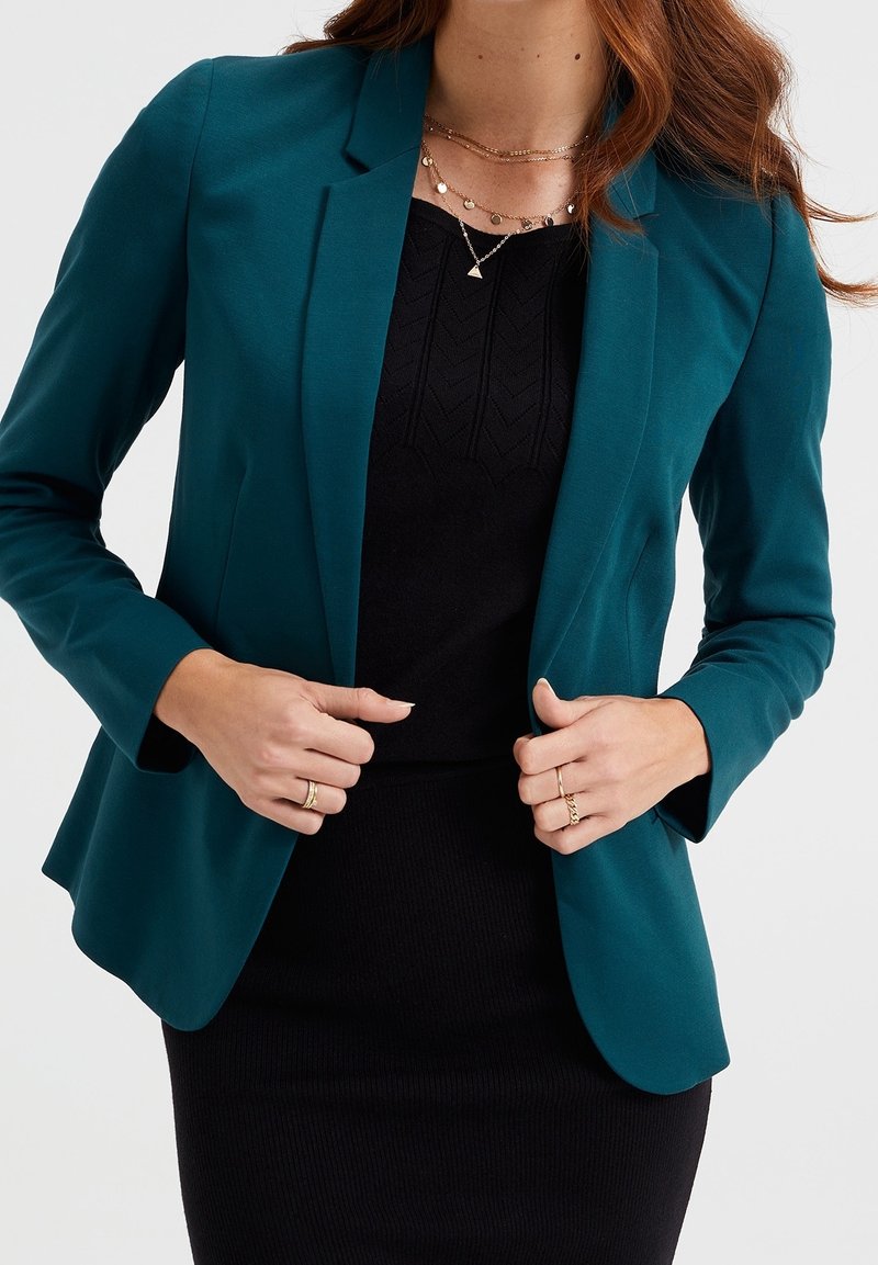 Femme ajustant un blazer bleu sarcelle sur une robe noire, portant des colliers en or superposés et des bagues aux deux mains.