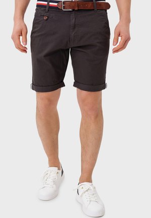 Man wearing rolled-up dark shorts, brown leather belt, a striped shirt tucked in, and white sneakers, standing against a plain background.