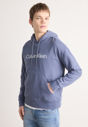 Sinine kapuutsiga soov, millel on eesmine tasku, nööridega kapuuts ja valge "Calvin Klein" logo. Sile kangas ja ribakujulised vöötohatised, vaba lõige.