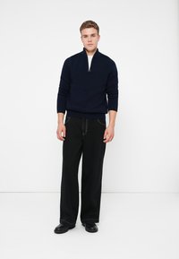 Maglione blu navy a maglia con colletto alto e chiusura con zip, abbinato a pantaloni neri a gamba larga e scarpe nere, su uno sfondo bianco semplice.