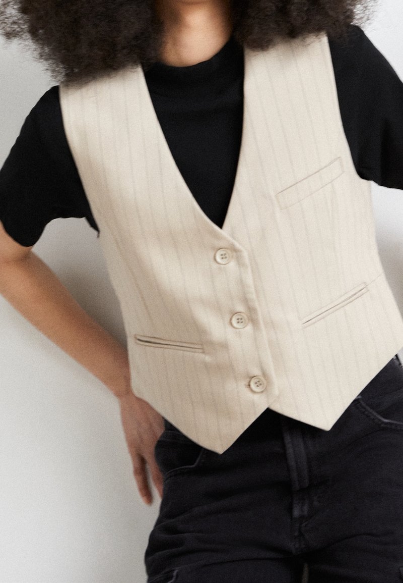 Personne portant un gilet beige à fines rayures sur un t-shirt noir, les mains sur les hanches devant un fond uni.