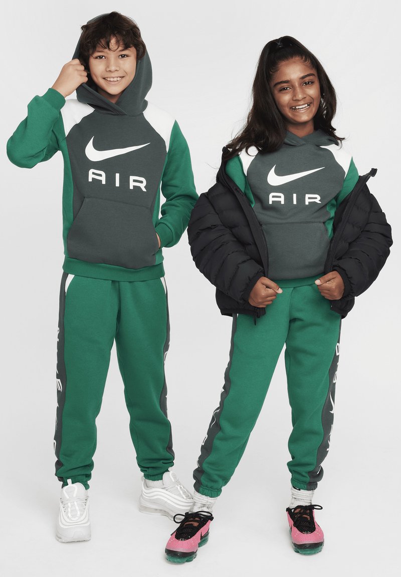 Dwoje dzieci nosi dopasowane, zielone dresy Nike z widocznym logo. Jedno z dzieci ma również na sobie czarną kurtkę puchową.