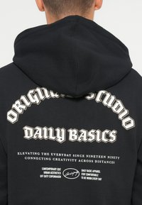 Hoodie noir avec un grand graphique blanc au dos, présentant "Original Studio" et "Daily Basics" en lettres stylisées et en gras.