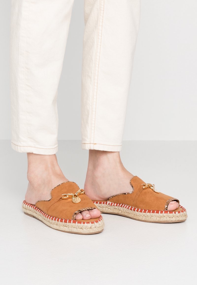 Pieds portant des sandales mule en daim beige à bout ouvert avec détail chaîne dorée et semelles intermédiaires en jute cousu, associées à un pantalon blanc cassé.