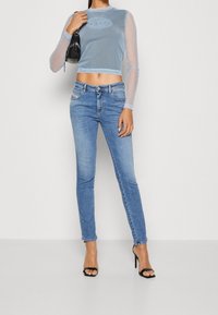Top de manga larga recortado en azul claro con mangas transparentes, combinado con unos vaqueros ajustados azules. Sandalias de tacón negro y un pequeño bolso negro completan el conjunto.