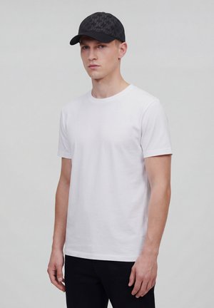 Ung mand iført en enkel hvid t-shirt med korte ærmer, sorte bukser og en sort baseballkasket med et diskret mønster, stående foran en grå baggrund.