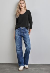 Schwarzer V-Ausschnitt-Pullover kombiniert mit blauen weit geschnittenen Jeans. Die Jeans haben einen verblassten Denim-Look und klassische Taschen. Weiße Sneakers runden den Look ab.