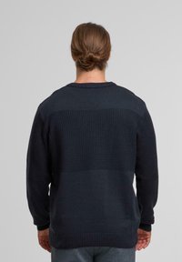 Persona con capelli castani legati, che indossa un maglione scuro a trama e pantaloni grigi, in piedi rivolto di spalle contro uno sfondo chiaro uniforme.