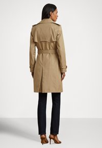 Καφέ χακί παλτό βροχής με ζώνη στη μέση, μακριά μανίκια, επιβραχιόνια και πλάτη με πιέτα· συνδυασμένο με σκούρα τζιν και καφέ ψηλοτάκουνες μπότες.