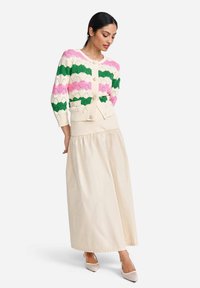 Strickjacke in Creme, Pink, Grün; geometrisches Muster; runder Halsausschnitt, Knopfschluss, Taschen. Fließender beiger Rock; knöchellang.