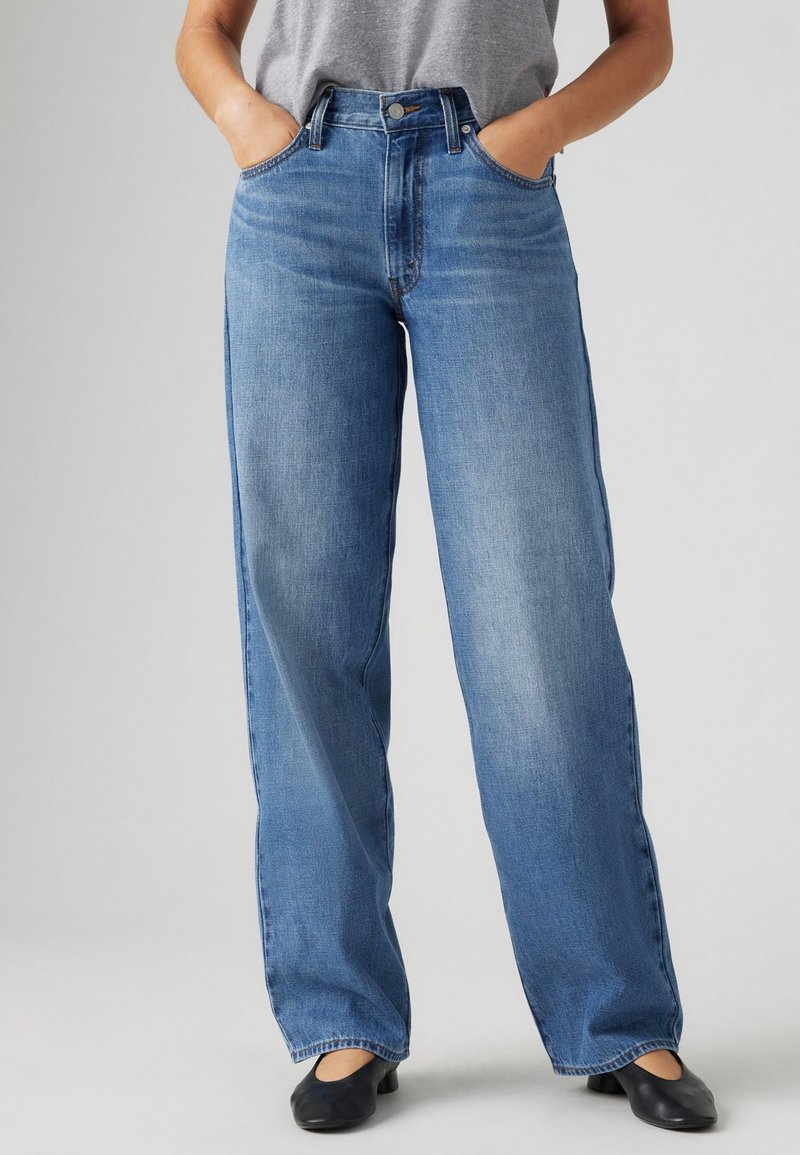 Personne portant un jean bleu taille haute, coupe ample, une chemise grise rentrée, des chaussures plates noires, debout les mains dans les poches devant un fond uni.