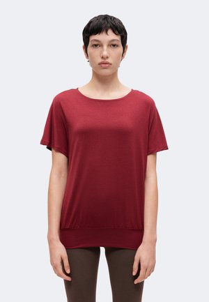 Persona con cabello corto y oscuro que lleva una camiseta roja lisa de manga corta y pantalones oscuros, de pie frente a un fondo gris claro.