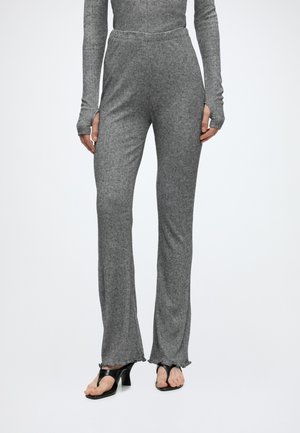 Personne portant un pantalon en maille côtelée gris avec ourlet volanté associé à des sandales noires à talons, montrée de la moitié du torse jusqu'aux pieds sur fond blanc.