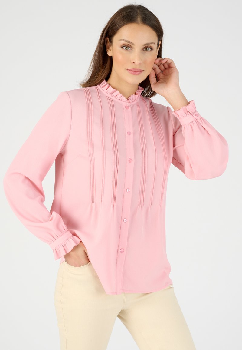 Blusa rosa con un cuello con volantes, detalles de pliegues verticales y cierre de botones en la parte frontal. Con mangas largas con puños elásticos y un corte holgado.