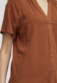 Camicia button-up color ruggine con scollo a V, maniche corte e un tessuto testurizzato, con cinque bottoni visibili nella parte anteriore.