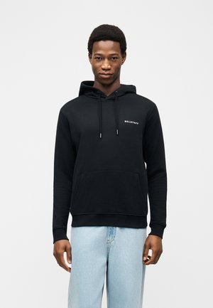 Junger Mann trägt einen schwarzen Belstaff-Hoodie und hellblaue Jeans, steht vor einem schlichten weißen Hintergrund.