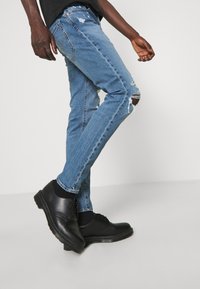 Slim-fit modré džíny s opotřebovanými detaily a roztrhnutím na koleni, zkombinované s černými koženými botami s hladkým povrchem a kulatou špičkou.
