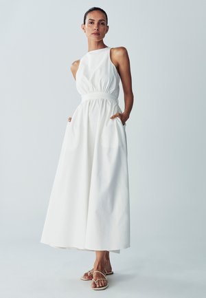 Weißes ärmelloses Maxikleid mit elastischem Bund und Seitentaschen, aus strukturiertem Stoff gefertigt. Das Modell trägt minimalistische Sandalen.
